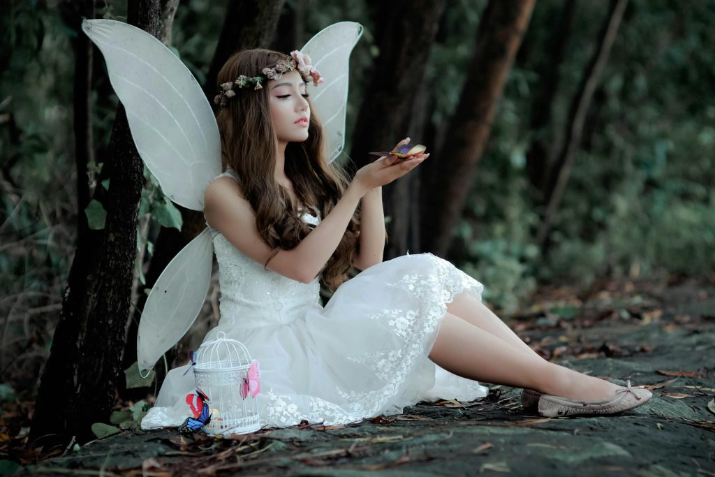 A captivating image of a fairy in white dress with wings, sitting in a woodland setting, holding a butterfly.
